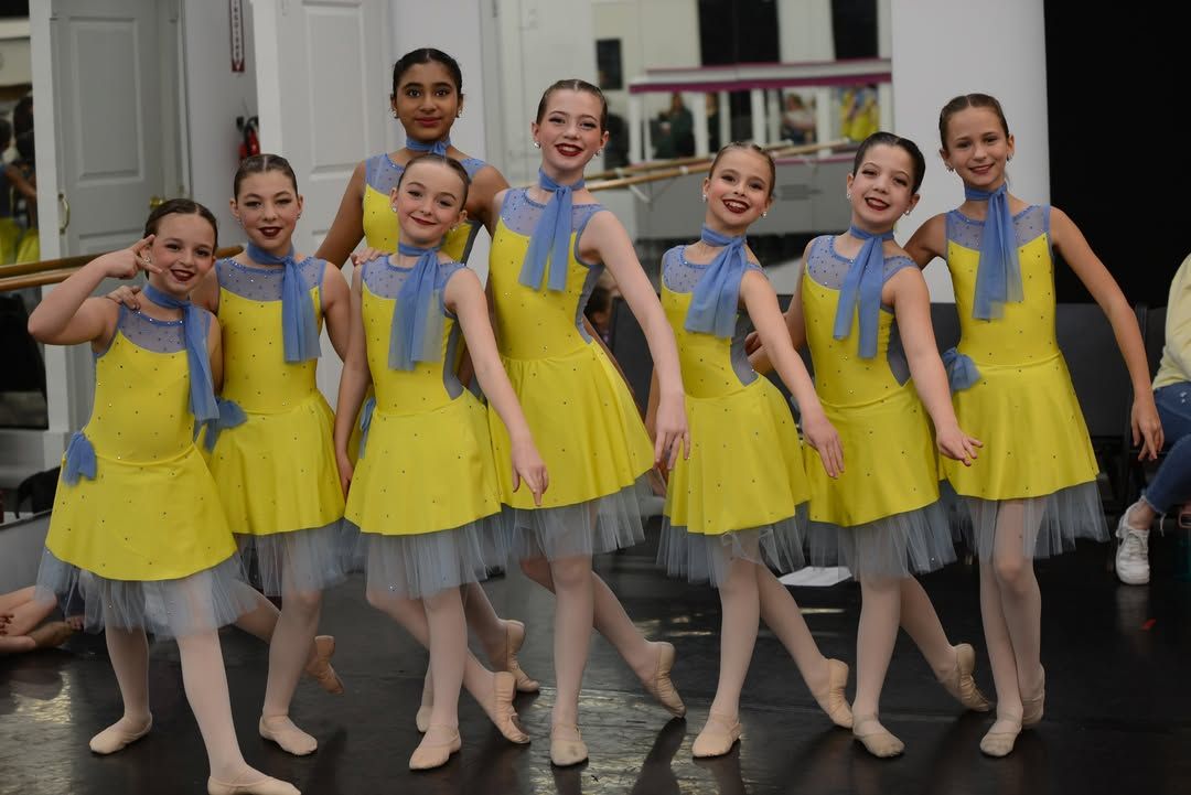 Group of dancers in yellow costumes posing on stage, smiling with arms linked