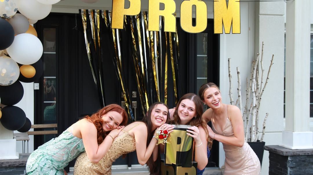 Four friends posing at a prom backdrop with gold “PROM” letters and black, white, and gold balloons