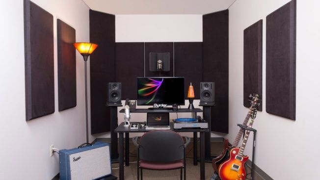 A room with a desk , chair , guitar , speakers and a computer.