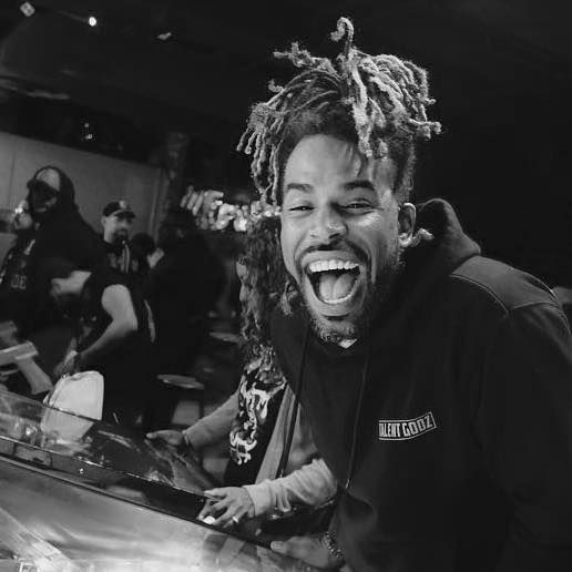 A man with dreadlocks is laughing with his mouth open in a black and white photo.