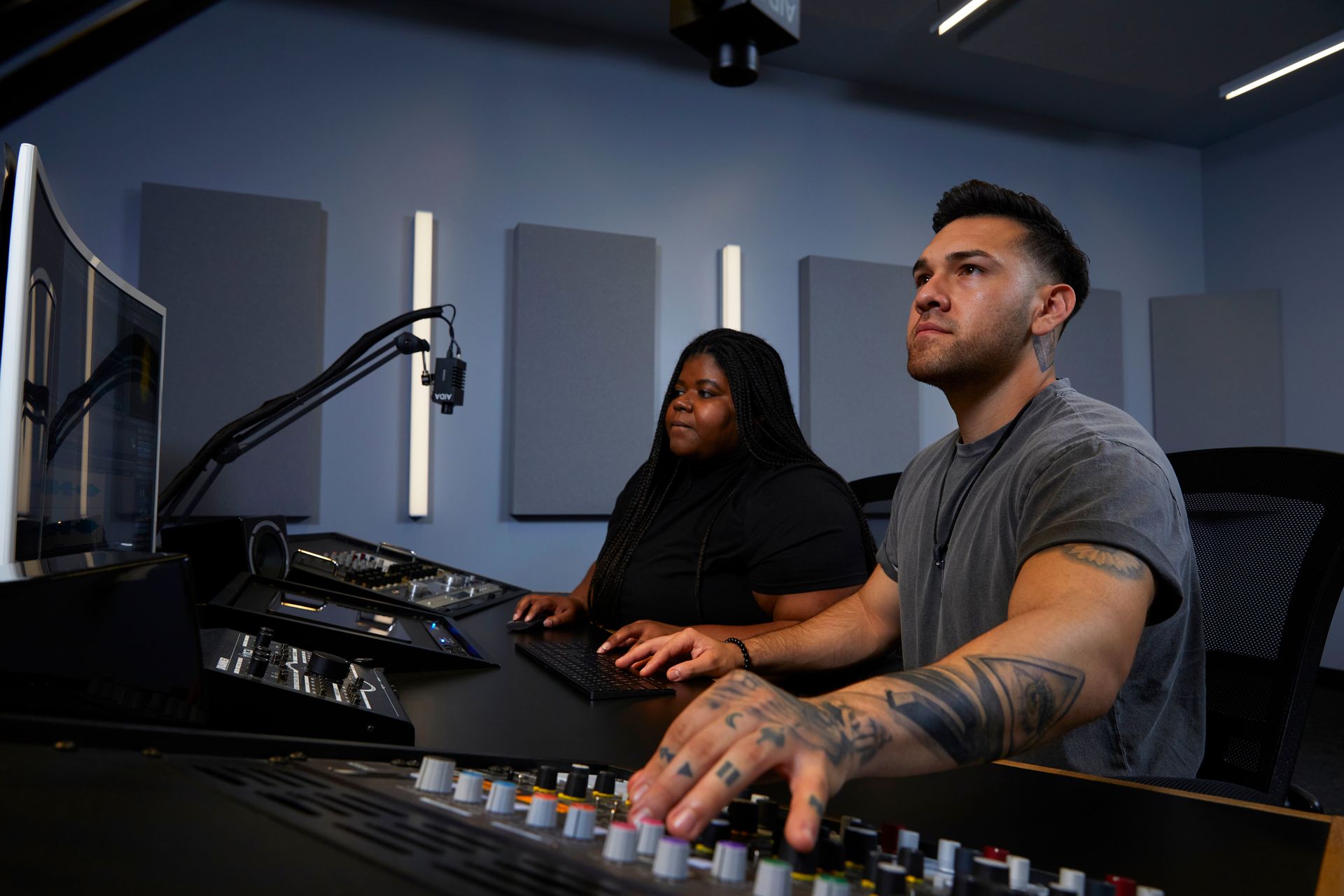 A man and a woman are sitting at a desk in a recording studio.