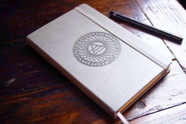 A notebook with a circle on the cover sits on a wooden table next to a pen