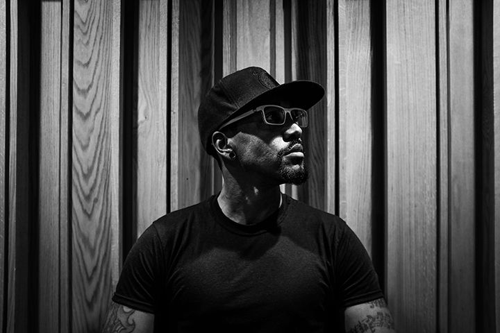 A man wearing a hat and sunglasses is standing in front of a wooden wall.