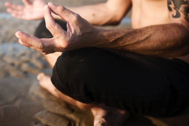 A man is sitting in a lotus position on the beach with his hands outstretched.