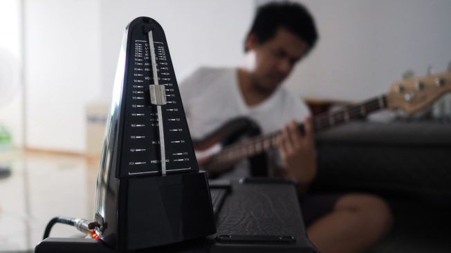 A man is playing a guitar with a metronome in the foreground.