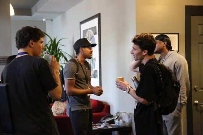 A group of young men are standing in a living room talking to each other.