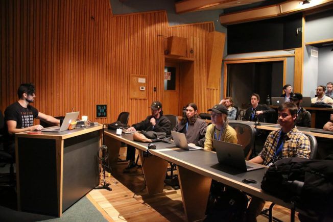 A group of people are sitting at tables with laptops in a classroom.