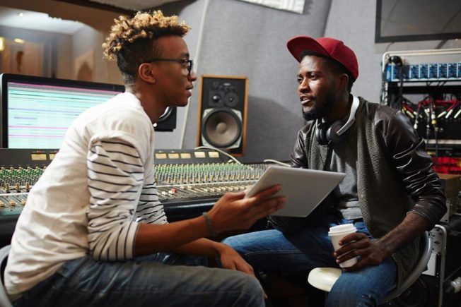 Two men are sitting in a recording studio looking at a tablet.