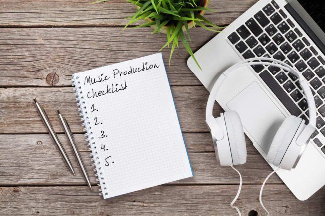 A music production checklist is written in a notebook next to a laptop and headphones