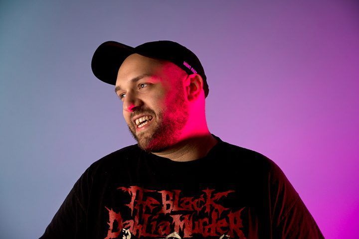 A man wearing a black t-shirt and a hat is standing in front of a purple background.