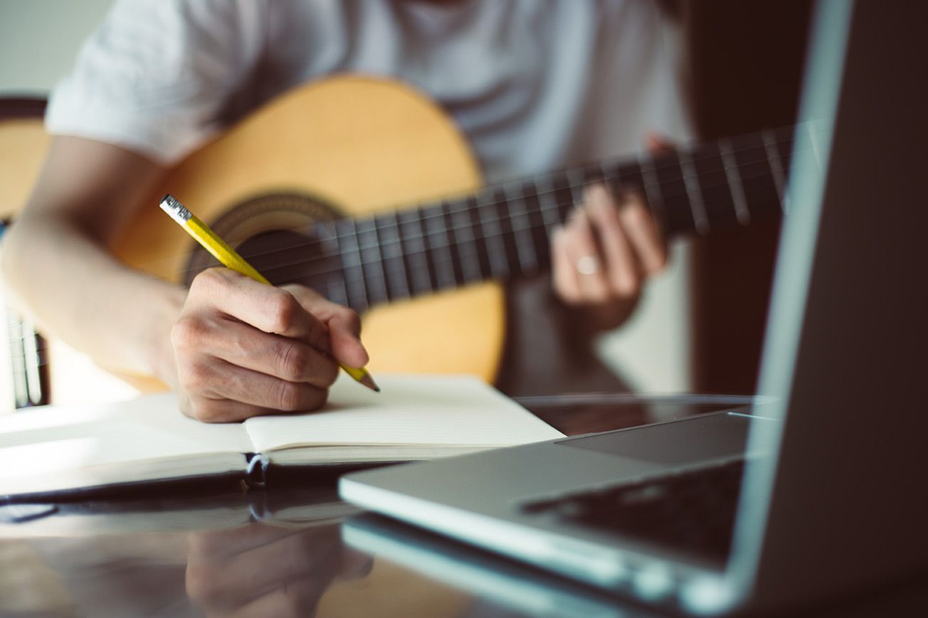 A man is writing in a notebook while playing a guitar.