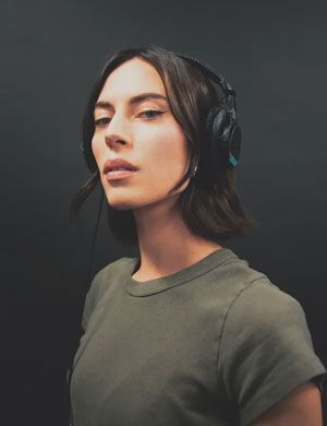 A woman wearing headphones and a green shirt is standing in front of a black wall.