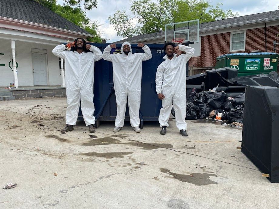 Three people in white coveralls pose with arms raised beside trash bins and a house driveway.