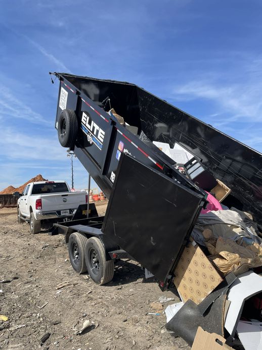 Damaged black dump trailer tipped steeply on rocky ground beside a white pickup truck under a blue sky