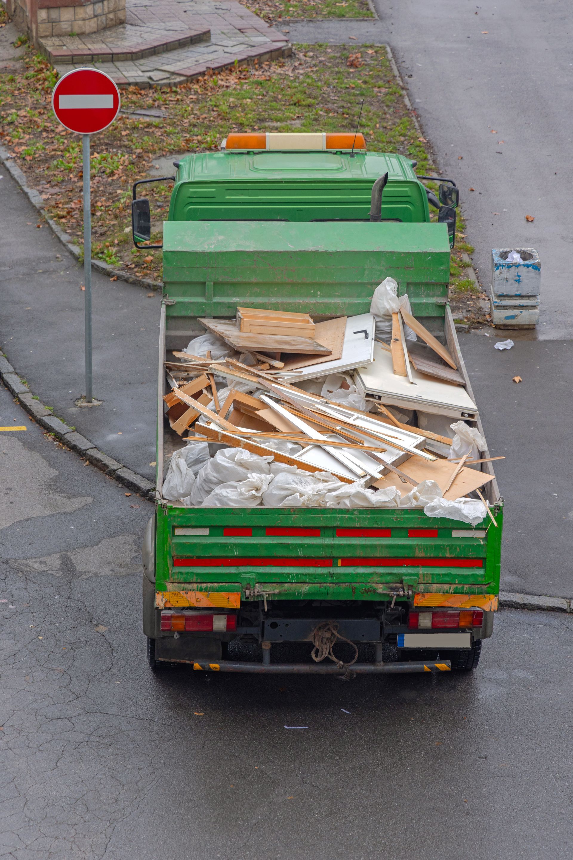 Green dump truck loaded with wood debris on a wet street near a no-entry sign