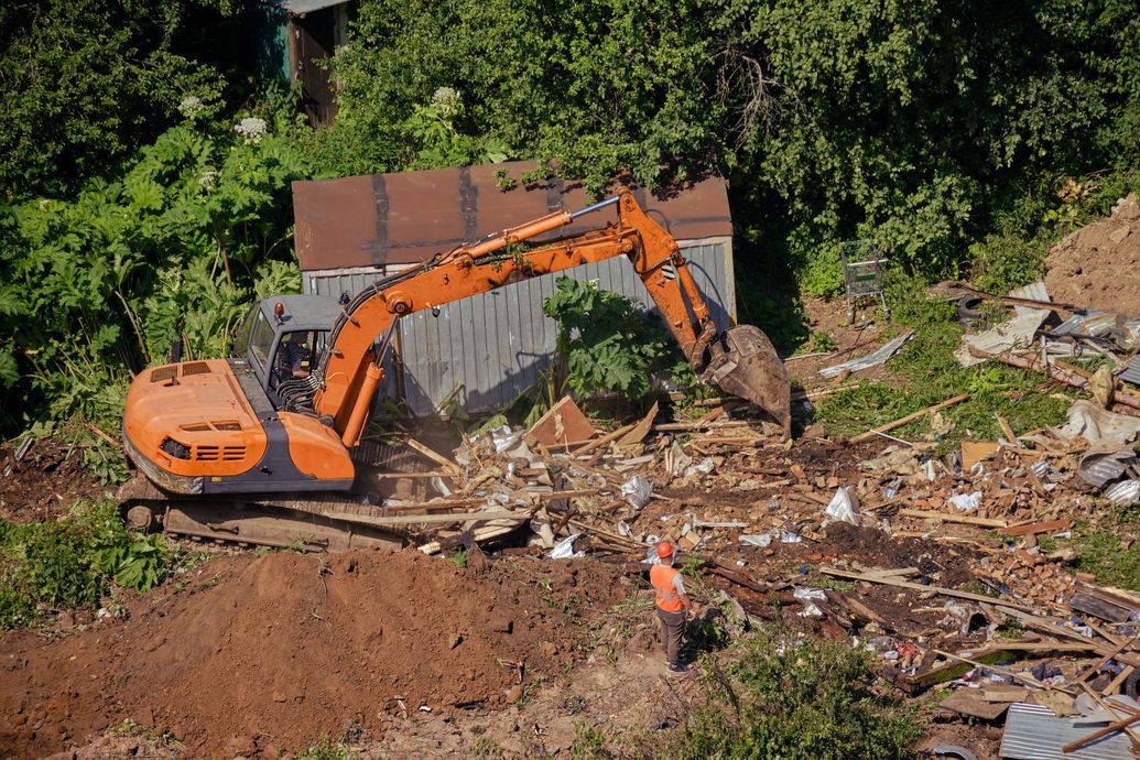 Orange excavator clearing debris in a wooded area near a damaged structure
