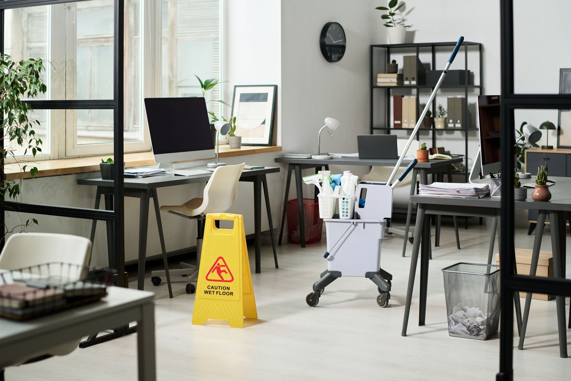 Modern office with desks, laptop, cleaning cart, and yellow wet floor sign near large windows