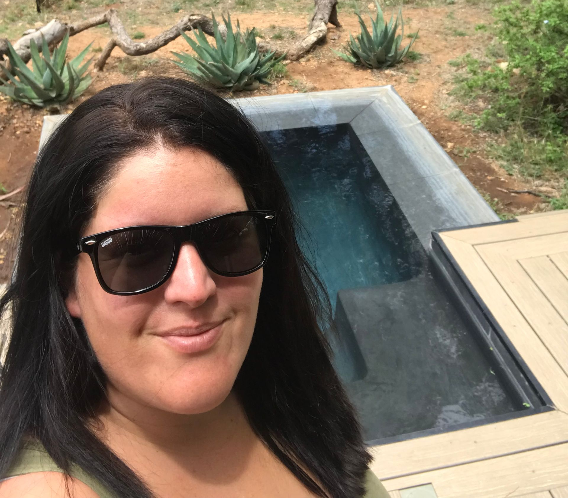 Woman in sunglasses smiles near a small, rectangular pool. Agave plants and a wooden deck are in the background.