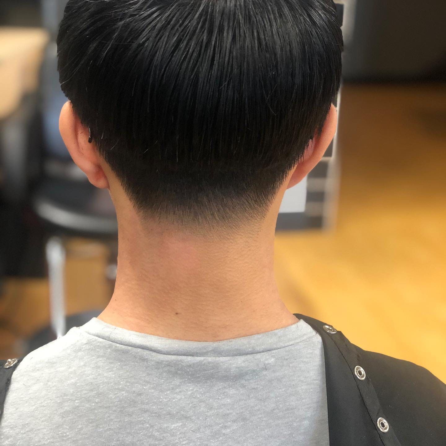 Back of a person with a short haircut in a barber chair, wearing a gray shirt in a salon.