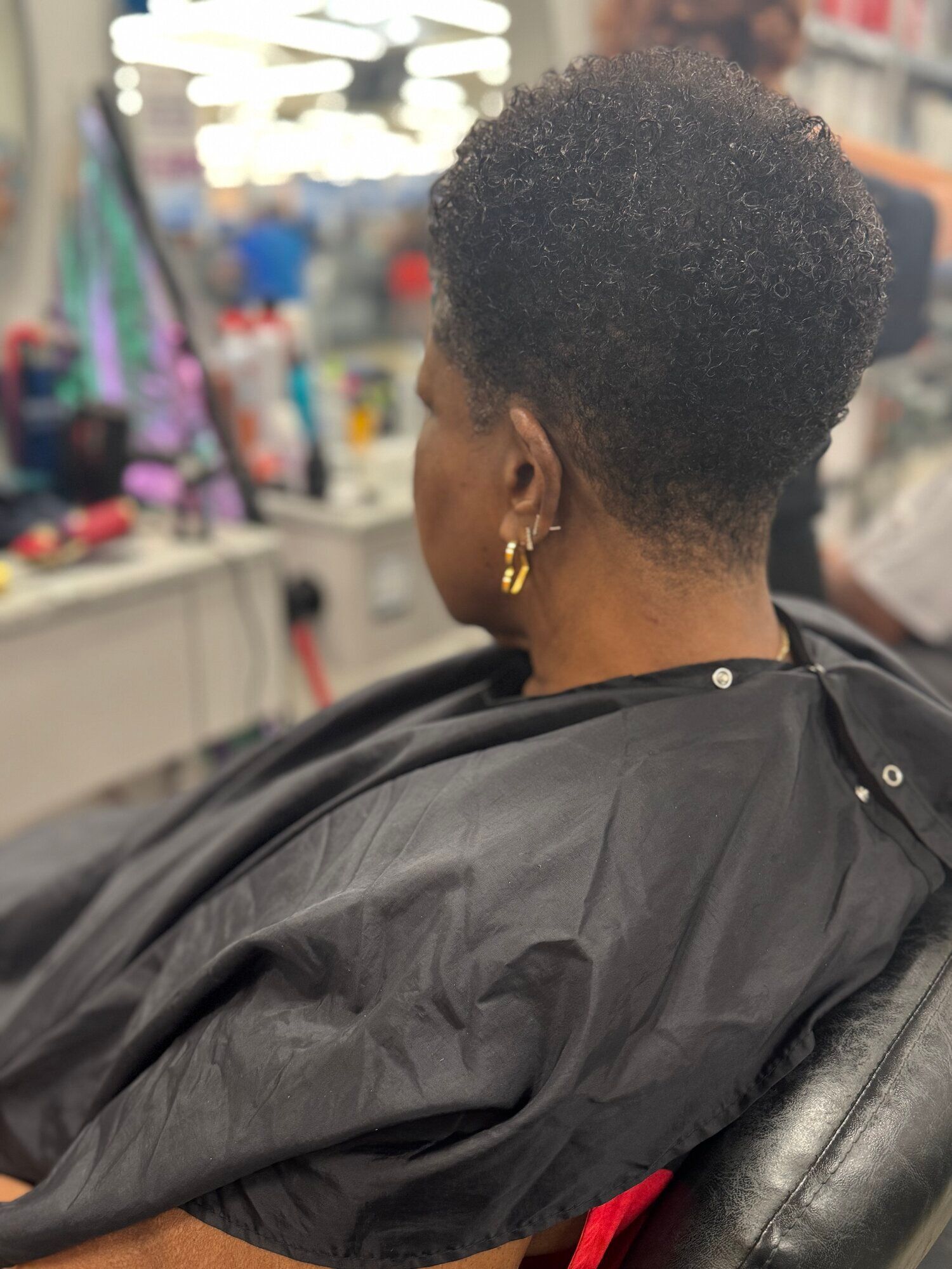 Person with short natural hair sitting in a salon chair, wearing a black cape, viewed from behind.