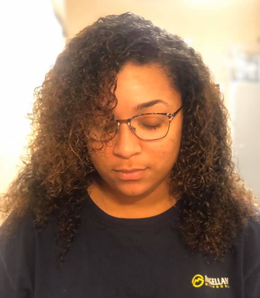 Person with curly hair and glasses looking down, wearing a dark shirt with a yellow logo.
