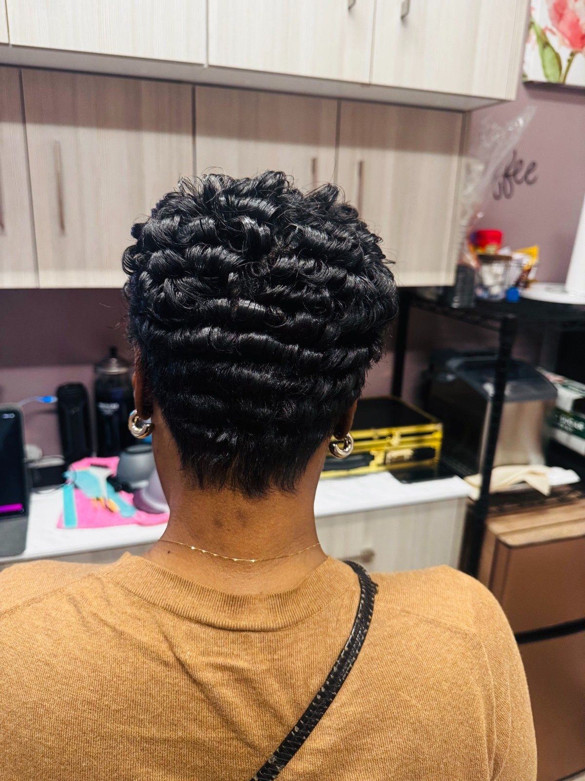 Short curly hairstyle viewed from behind in a salon chair, with a tan sweater and kitchen cabinets in the background