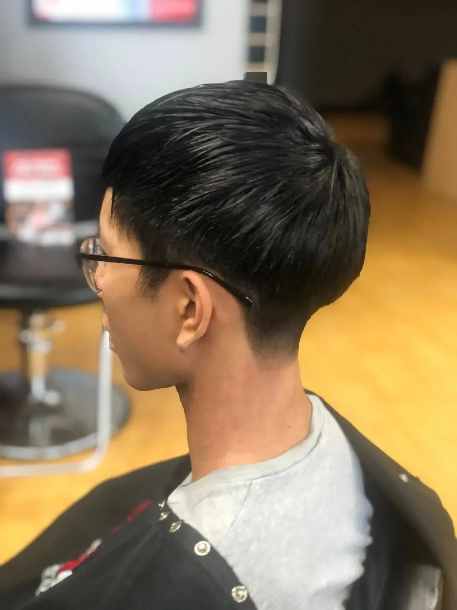 Side view of a person with short black hair and glasses in a salon chair, wearing a gray shirt.