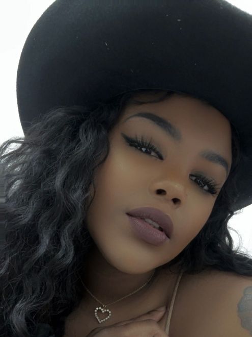 Close-up portrait of a woman wearing a black hat, with wavy hair and bold makeup.