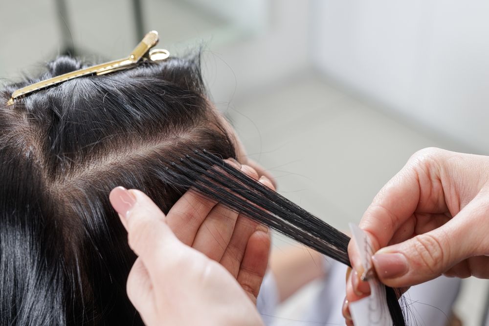 Hair extensions being attached to dark hair with a gold clip and hands sectioning strands