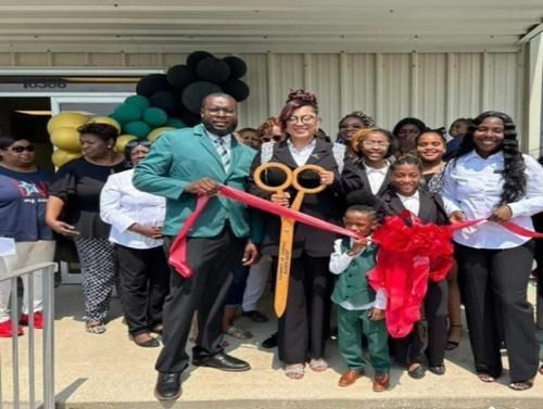 A group of people gathers for a ribbon-cutting ceremony outside a building decorated with a green and yellow balloon arch.