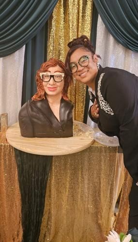 A smiling person stands next to a sculpted cake shaped like their own likeness, both wearing similar black-rimmed glasses.