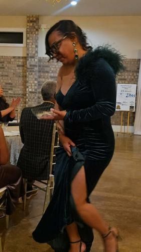 A person in a dark green velvet, one-shoulder dress with a high slit, standing in a room with tables and chairs.