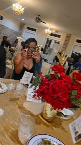 A person in a black dress holds a phone up to take a photo at a table decorated with a large bouquet of red roses.