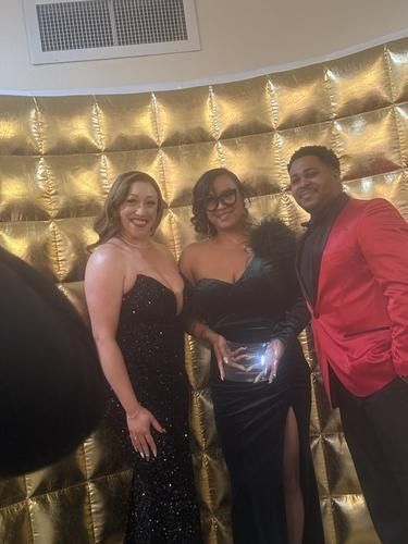 Three people smiling in formal wear, standing before a reflective gold backdrop. One person holds a clear trophy.