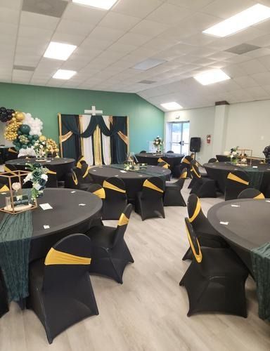 A dining hall set for an event with round black tables, chairs decorated with gold sashes, and a draped green-gold backdrop.