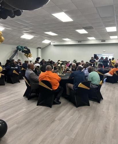 People gathered at round tables in a large event hall decorated with black and gold balloons for a formal event.