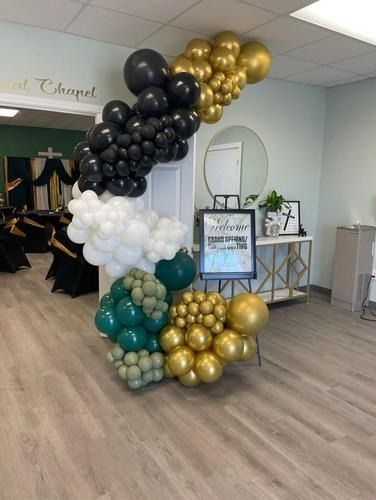 An arch of balloons in black, white, gold, green, and sage next to an easel sign in a brightly lit event space.