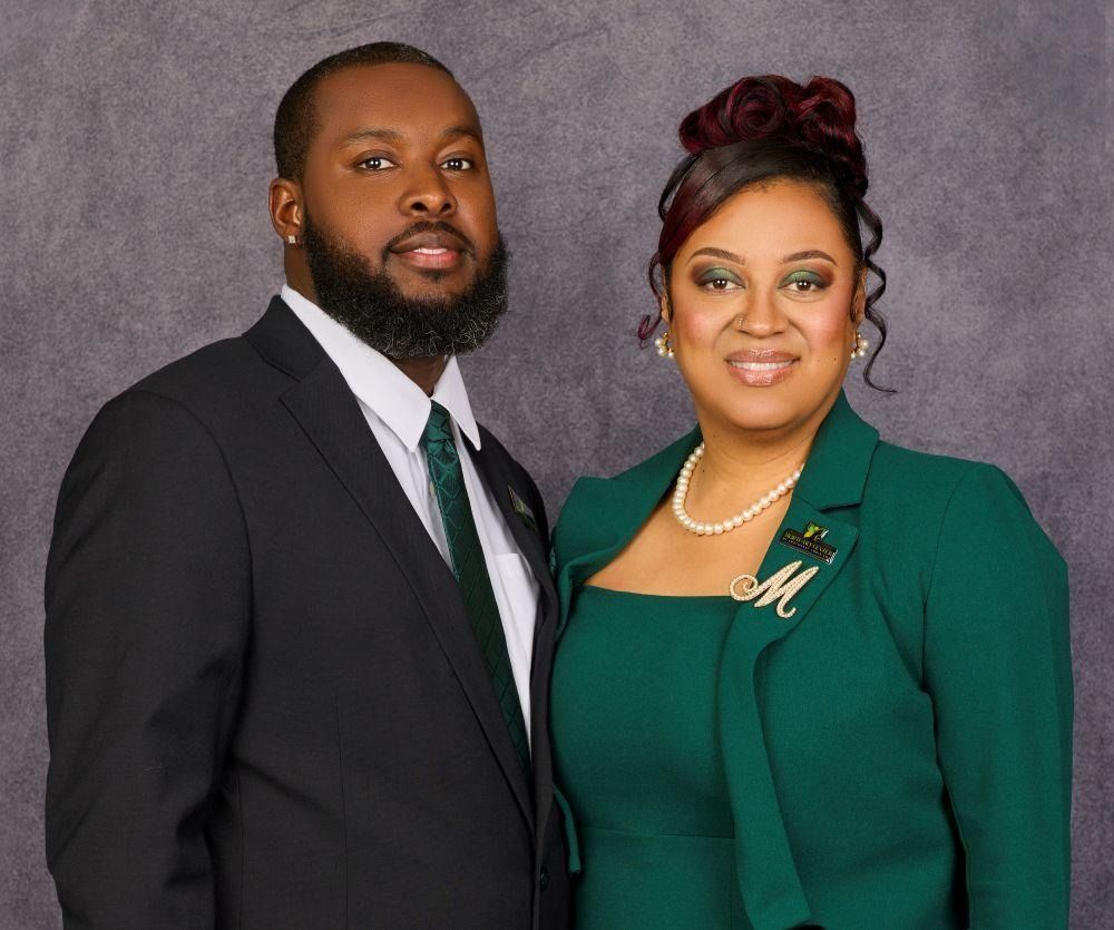 A man in a dark suit and green tie poses next to a woman in a green blazer and pearl necklace against a grey background.