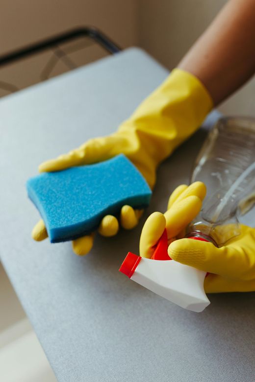 Hands in yellow gloves holding a blue sponge and a spray bottle on a table.