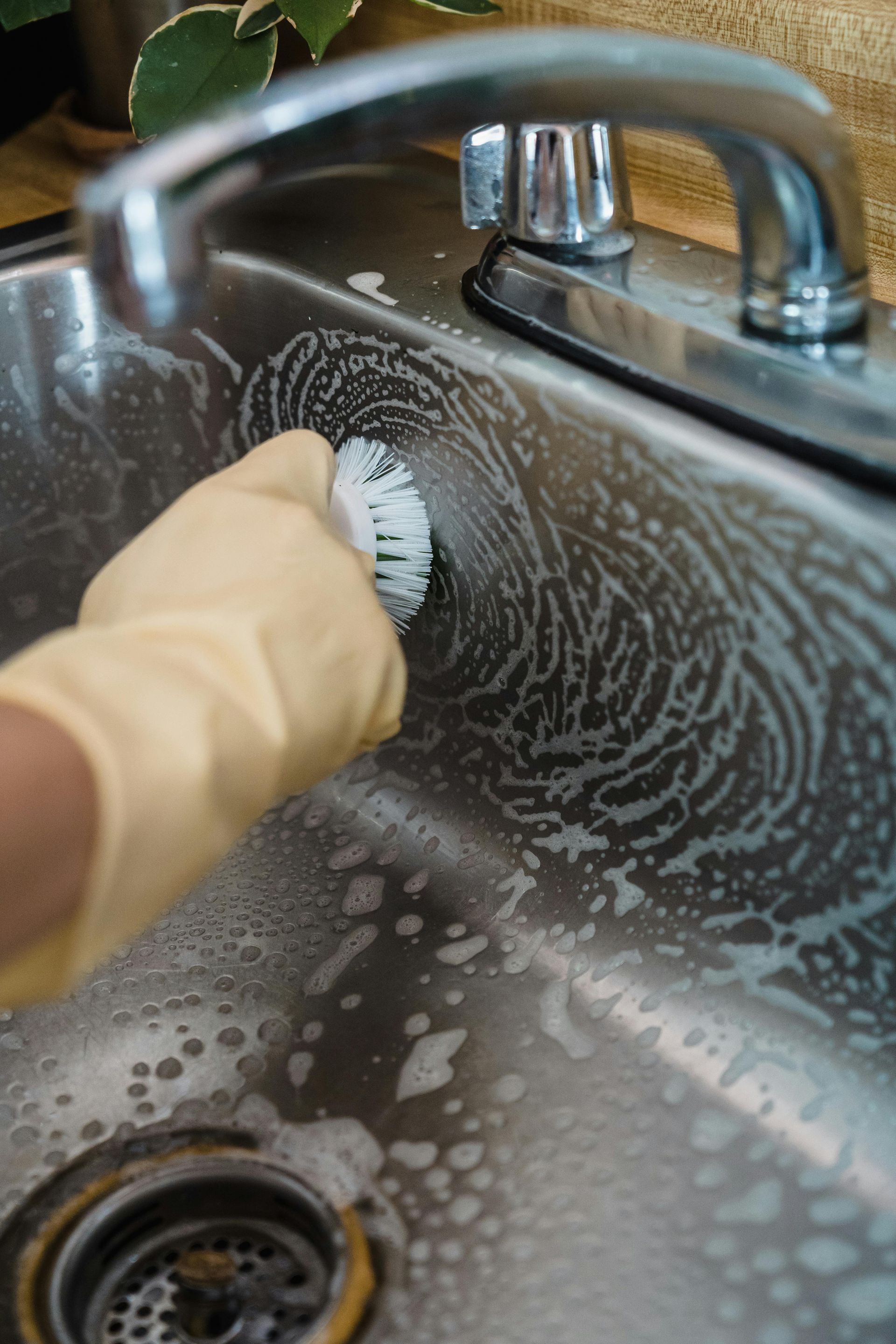 A person wearing a glove scrubs a stainless steel sink with a brush and soapy water.