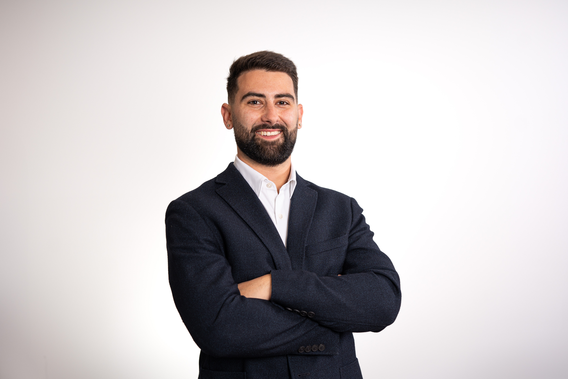 Man with beard, wearing navy blazer and white shirt, smiling in front of a white backdrop.