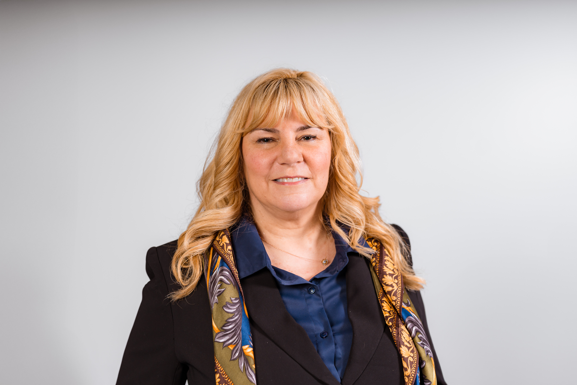 Woman with blonde hair wearing a patterned top and black blazer, smiling with one hand on hip.