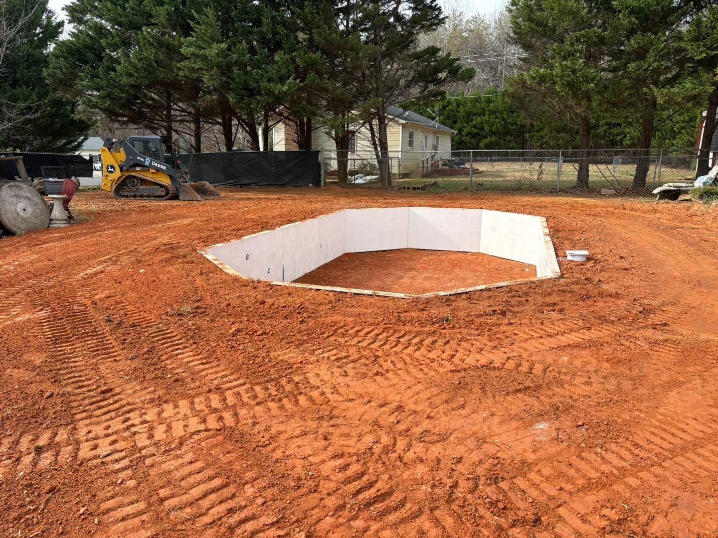 Pool excavation in progress: bare earth surrounding a white-walled, geometric pool form. Yellow construction vehicle in background.