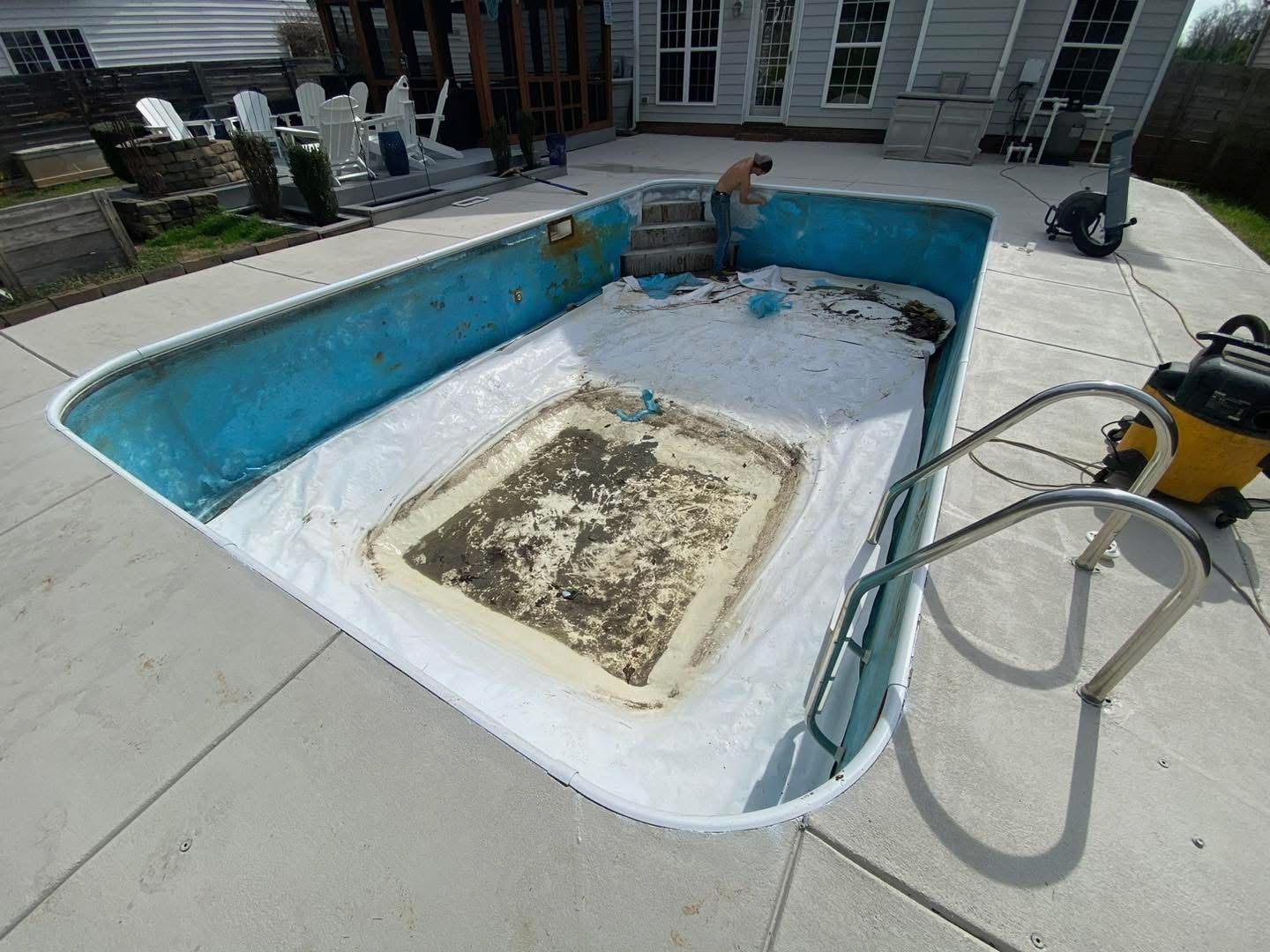 Pool being cleaned. A person in the pool with a vacuum. Concrete surrounding with a house in the background.