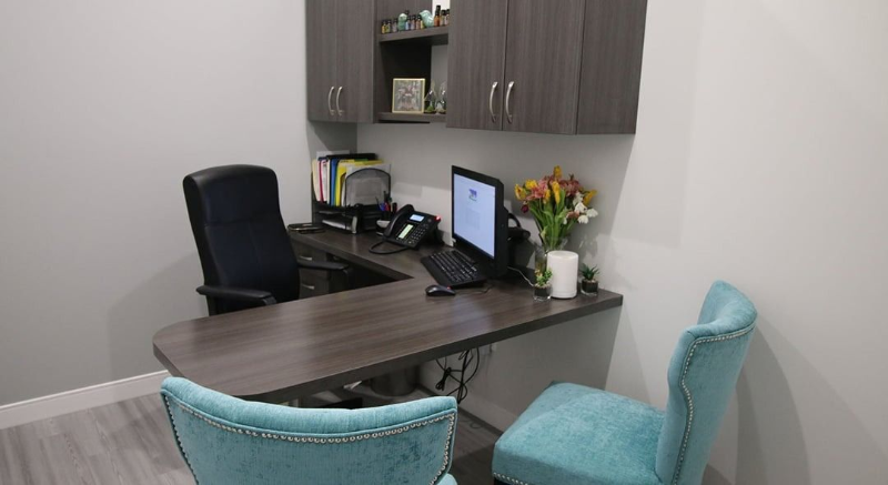 Small office with a desk, computer, black chair, and two teal chairs beside a wall cabinet.
