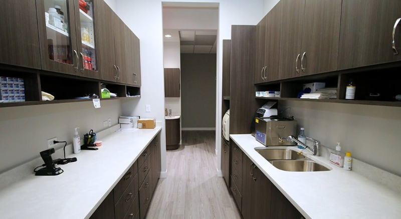 Modern office kitchenette with two rows of dark wood cabinets and white counters, viewed down a narrow aisle