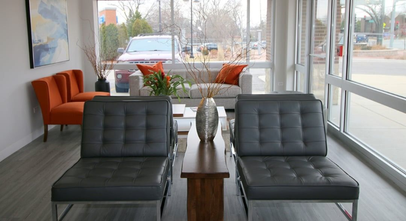 Modern lobby seating area with black lounge chairs, orange armchairs, and a glass table by large windows
