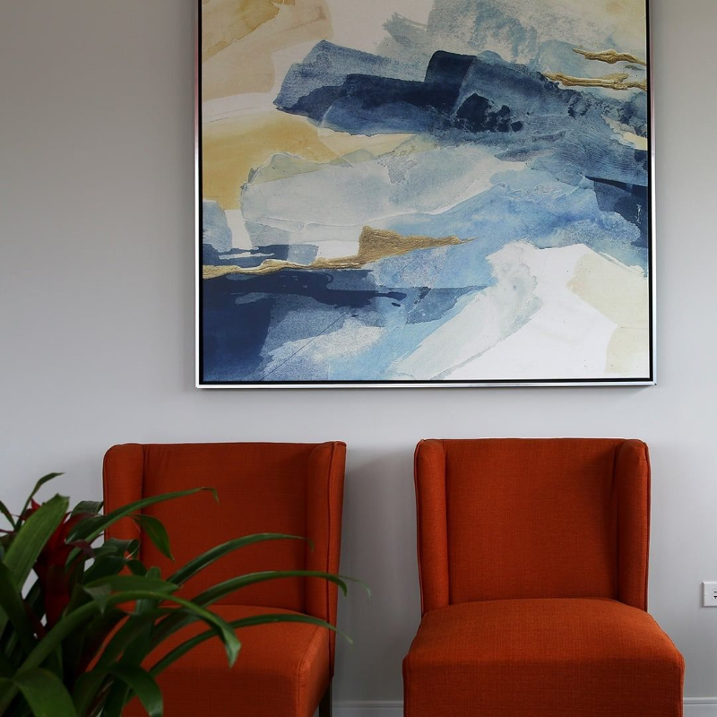Two orange chairs beneath an abstract blue and cream painting, with a green plant in the foreground.