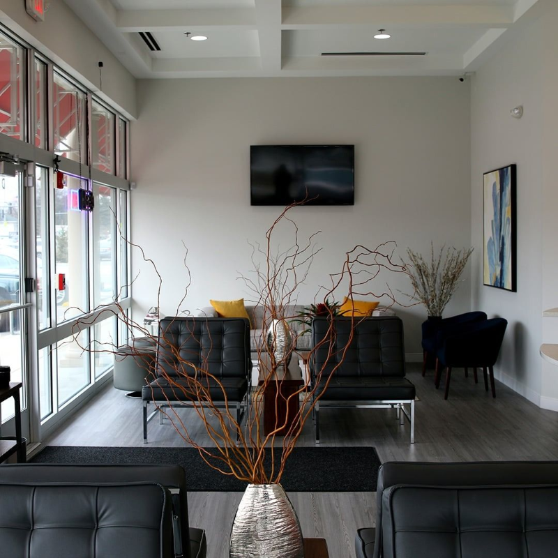 Modern lobby with black chairs, wall-mounted TV, and a vase of branches on a table; glass doors on the left