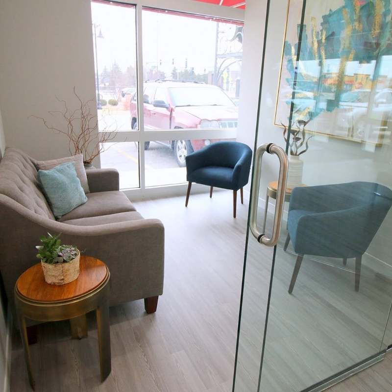 Bright waiting room with gray sofa, blue chairs, glass door, and round wooden table by a window